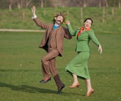 Still from film. Two characters in period costume skipping through field.